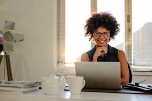 Portrait of smiling young woman sitting at her desk with laptop. African female sitting at a table with laptop looking at camera and smiling in office.